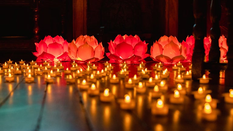 Orange glowing candles are arranged in circles on a dark wood floor with pink lit lotus flower displays in the background